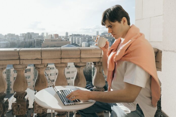 Mężczyzna pracuje przy laptopie i pije kawę na miejskim balkonie Mężczyzna pracuje przy laptopie i pije kawę na miejskim balkonie